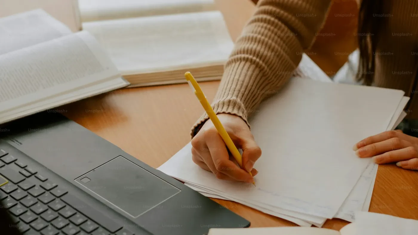 Student writing beside a laptop and open book